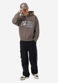 Brauner Kapuzenpullover mit Reißverschluss, vorn Taschen und grafischem Text, kombiniert mit schwarzen Cargohosen und einer passenden Mütze mit ähnlichem Schriftzug. Sneakers vervollständigen das Outfit.