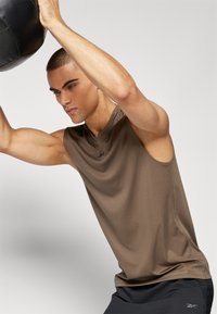 Camiseta sin mangas de color marrón con textura suave, con cuello redondo y detalles de costura mínimos, llevada por un modelo masculino.