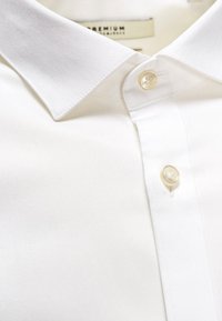 Camicia bianca con colletto, realizzata in tessuto liscio e morbido, dotata di una chiusura a un singolo bottone e di una leggera lucentezza sulla stoffa.