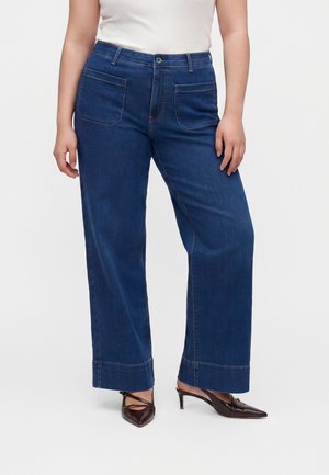Kvinde iført vidde mørkeblå jeans med forlommer og brune spidse hælede sko mod en ensfarvet baggrund.