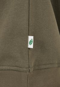 Tessuto di felpa verde oliva con un motivo a maglia testurizzata, caratterizzato da un'etichetta bianca con logo a foglia verde all'orlo e polsini a costine.