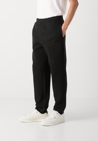 Pantalons de survêtement noirs en matériau doux, avec une taille élastique, des poches latérales et un petit logo sur la cuisse. Associés à des baskets blanches.