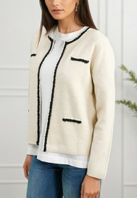 Veste en mélange de laine ivoire avec des finitions noires, col rond, et deux petites poches poitrine, portée sur un haut blanc, associée à un jean bleu.