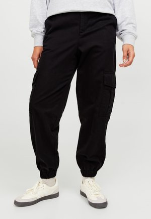 Pantalons cargo noirs avec des poignets élastiques, de multiples poches sur les côtés et une coupe décontractée, fabriqués en tissu durable, associés à des baskets blanches.
