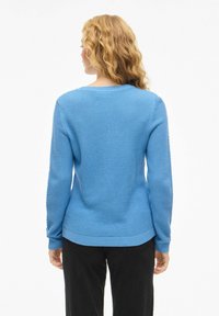 Niebieski sweter dziany z fakturowanym wzorem, okrągłym dekoltem i ściągaczami na rękawach. Noszony z czarnymi spodniami, prezentujący swobodny widok od tyłu.