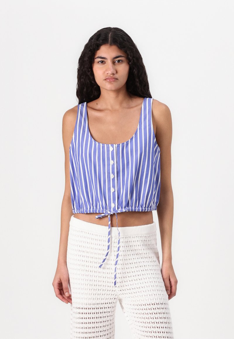 Top corto a righe blu e bianche con scollatura a U, bottoni sul davanti e laccio regolabile all'orlo. Indossato con pantaloni bianchi testurizzati.