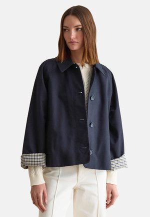 Mujer con cabello castaño liso, vistiendo una chaqueta azul marino con puños a cuadros, suéter crema y pantalones crema, mirando hacia la izquierda sobre un fondo liso.