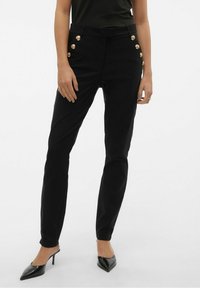 Pantalones negros con acentos de botones dorados en un lado, diseño ajustado y una textura suave. Llevados con tacones negros de punta.