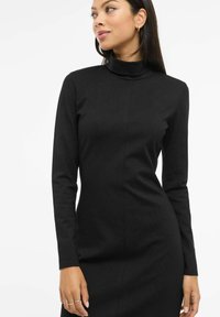 Černé šaty s dlouhými rukávy z hladké látky, s vysokým límcem a přiléhavým střihem, s švovým zdobením na přední straně.