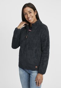Black, fuzzy fleece jacket with a zip-up front, high collar, two side pockets, and logo accent on the lower hem. Soft texture and warm design.
