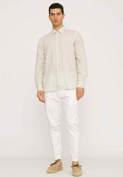 Camicia in lino button-up di un beige pallido con colletto e maniche lunghe, indossata con pantaloni bianchi e scarpe marroni chiare. Texture liscia, design minimal.