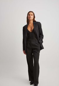 Una donna indossa un top in pizzo nero con corsetto sotto un blazer nero oversize, abbinato a pantaloni a vita alta neri e scarpe nere con punta.