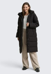 Zwarte pufferjas met een lange, gewatteerde design, voorzien van een capuchon, een rits aan de voorkant en drukknopen. Het model draagt beige broek en laarzen.