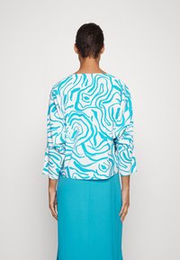 Blusa a maniche corte in tessuto bianco con motivi astratti blu a spirale, abbinata a una gonna turchese uniforme.