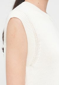 Tricou fără mâneci tricotat, de culoare albă murdară, cu un guler rotund ribat și cusături pe umeri. Include detalii cu deschideri în apropierea brațelor.