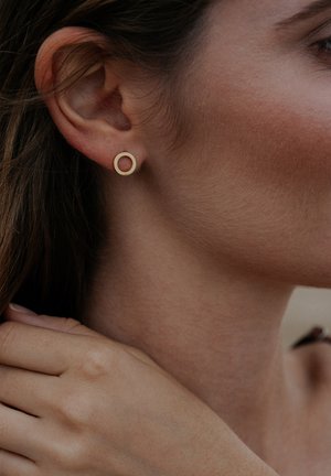 Cercel cu tărtăcuță circulară aurie, design minimalist, textură mată, așezat într-o fotografie în prim-plan a unei urechi, cu un ton al pielii neutru ca fundal.