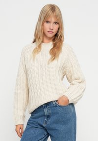 Cream knitted sweater with a ribbed texture, wide sleeves, and a high neckline, paired with light blue denim jeans.