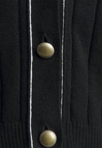 Svart stickad cardigan med vertikal ribbad textur, vit kontrastsömnad och stora runda bronseknappar på framsidan.