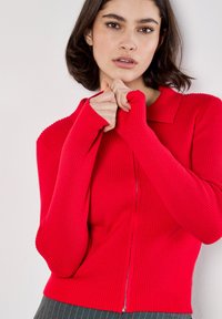 Czerwony sweter ze żebrowanym wzorem, z kołnierzykiem, długimi rękawami i zamkiem błyskawicznym z przodu. Charakteryzuje się teksturowanym materiałem i dopasowanym krojem.