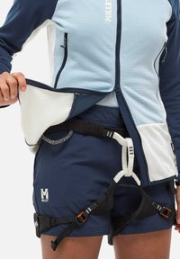 Person wearing blue and white Millet jacket and navy climbing shorts with a climbing harness secured around their waist and legs.