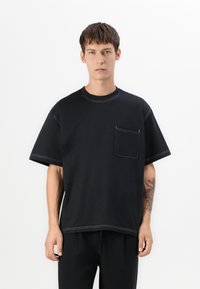Svart t-skjorte med kortermet i bomull, med brystlomme, kontraststing og en avslappet passform. Glatt tekstur med minimalistisk design.