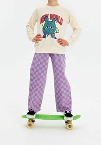 Kremowa bluza z kolorową grafiką, zestawiona z lawendowymi spodniami w kratę o szerokich nogawkach. Stojąc na zielonej deskorolce w czarnych butach.