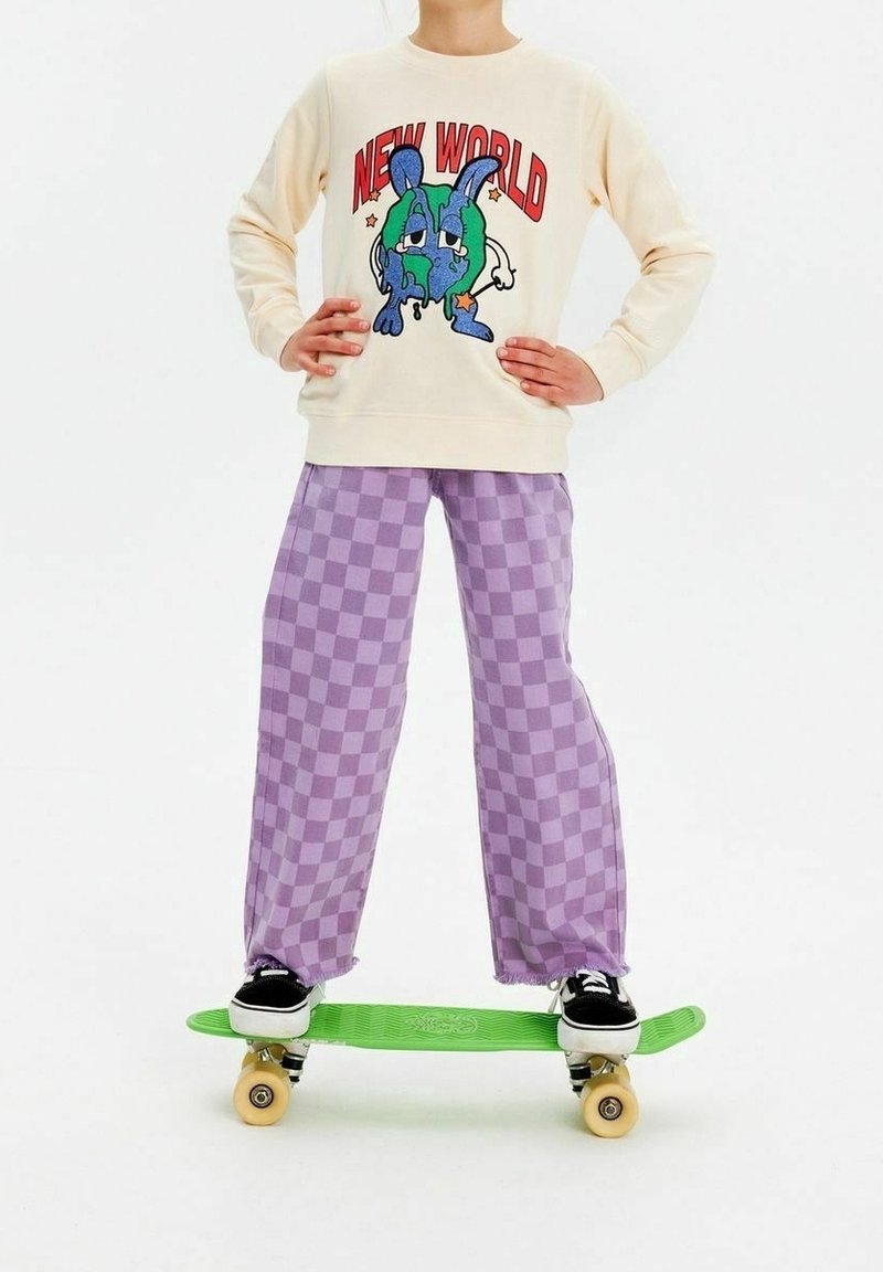 Kremowa bluza z kolorową grafiką, zestawiona z lawendowymi spodniami w kratę o szerokich nogawkach. Stojąc na zielonej deskorolce w czarnych butach.