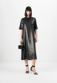 Donna che indossa un vestito midi nero lucido con maniche corte, tiene una piccola borsa nera e indossa sandali neri con tacco alto.