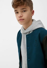 Teal wollen varsity jack met zwarte leren mouwen en drukknoopsluitingen, gedragen over een grijze sweatshirt met een capuchon en gestreepte accenten.