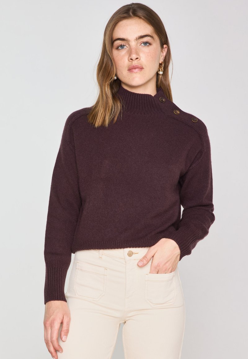 Pull en tricot bordeaux avec un col haut et des boutons sur les épaules, associé à un pantalon beige clair avec des poches avant.