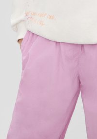 Weiche, rosa Pants mit lockerer Passform, elastischem Bund, Seiten-Taschen und glatter Textur. Teilweise von einem weißen Sweatshirt mit orangefarbigem Text verdeckt.
