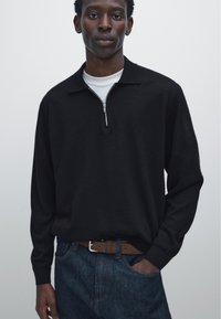 Young man wearing a black zip-up sweater over a white shirt tucked into blue jeans with a brown belt, standing against a plain background.