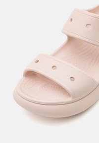 Sandal roz deschis, fabricată din cauciuc texturat, având două bretelelate cu găuri circulare și o talpă căptușită. Talpa rotunjită pentru confort.