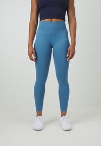 Leggings blu a vita alta realizzati in tessuto elastico, con una texture liscia e un design aderente, abbinati a scarpe da ginnastica bianche.