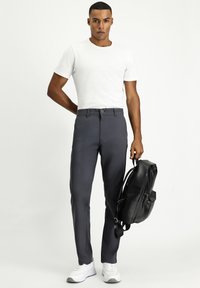 Grijze op maat gemaakte broeken met een knoopsluiting, gecombineerd met een witte t-shirt met korte mouwen. Model houdt een zwarte rugzak vast. Witte sneakers.