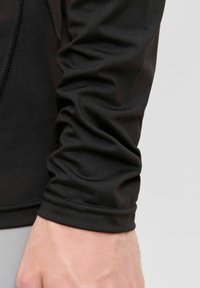 Ärmel eines schwarzen, taillierten Kleidungsstücks mit sichtbaren Nähten, leicht zerknittert um das Handgelenk, das über einer hellen Hand liegt.