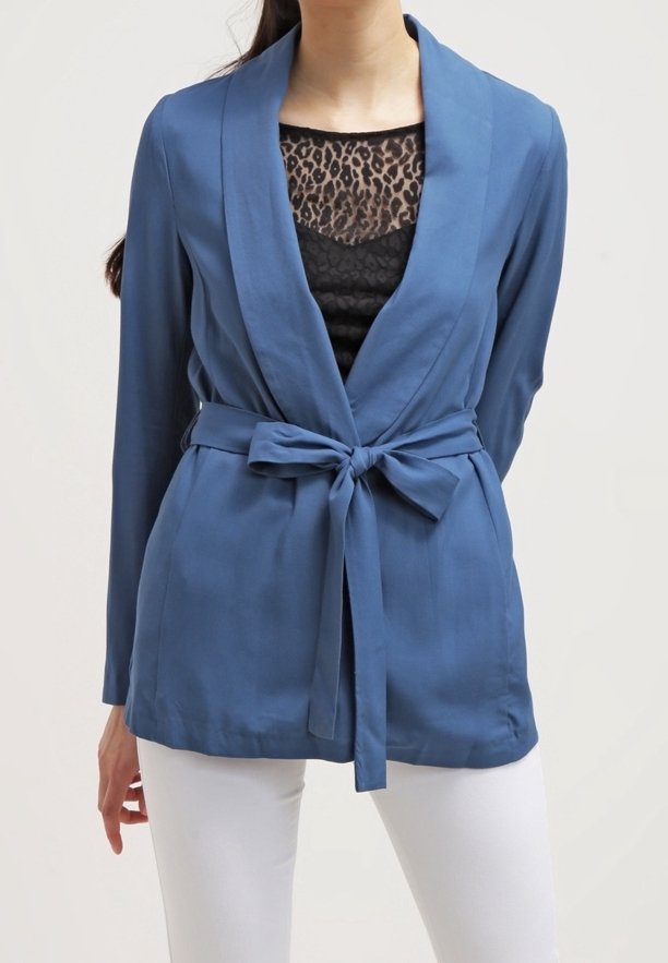 Femme portant un blazer bleu ceinturé sur un haut à imprimé léopard noir et un pantalon blanc, debout les mains derrière le dos devant un fond uni.