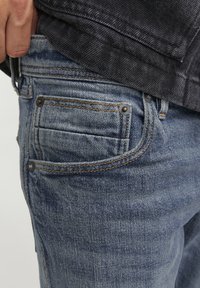 Jeans din denim într-o nuanță deschisă de albastru, cu un material texturat. Caracteristicile includ cusături portocalii, accesorii de culoare închisă și un design vizibil al buzunarului din spate.