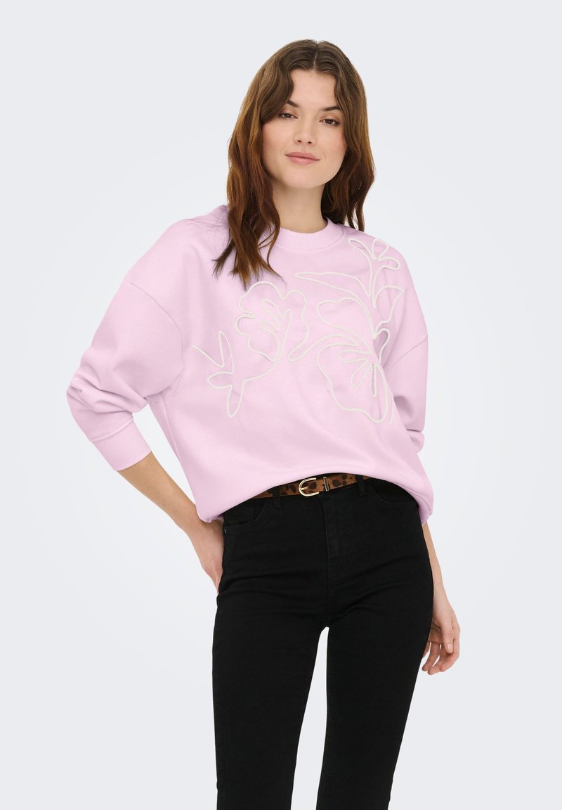 Jeune femme aux longs cheveux bruns portant un sweat-shirt rose clair avec broderies florales, un pantalon noir et une ceinture à imprimé léopard, posant avec une main en arrière.