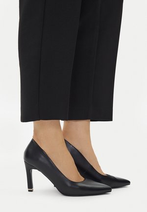 Füße in schwarzen spitzen High Heels kombiniert mit schwarzen, hochgekrempelten Hosen vor einem schlichten hellen Hintergrund.