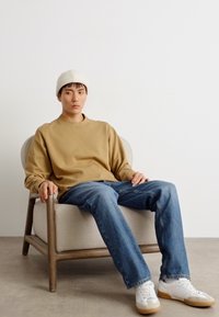 Sudadera beige de cuello redondo, jeans de mezclilla azules, zapatillas blancas con suelas de goma. Modelo sentado en una silla de madera contra un fondo simple.