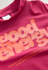 Fuchsia kortærmet T-shirt lavet af glat stof med fed, orange typografi, der indeholder sætningen "god energi" og yderligere tekst.