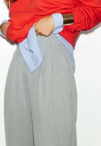 Un pantalon gris à rayures avec des plis, associé à un sweat-shirt rouge. Une chemise bleu clair est rentrée à l'intérieur et un bracelet à frapper en or est visible.