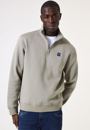 Lichtgrijze half-zip sweatshirt van zacht materiaal, met een klein blauw logo op de borst en geribbelde boorden en zoom. Draag over een wit shirt.