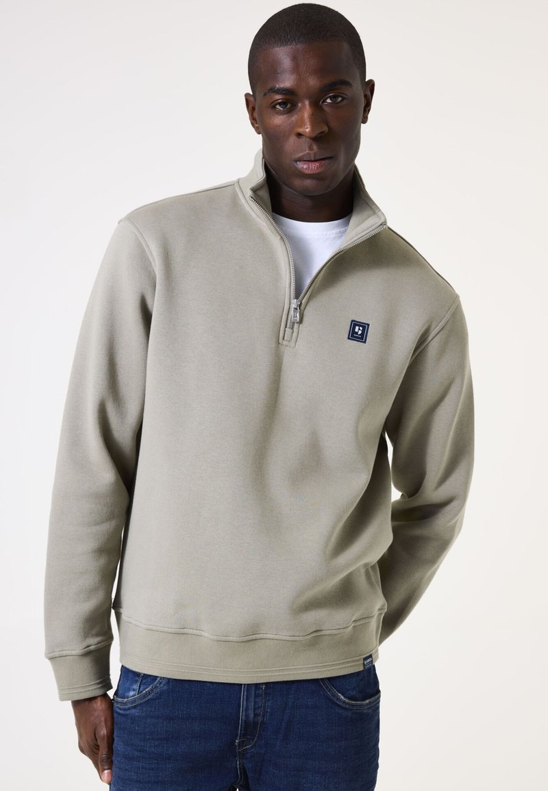 Lichtgrijze half-zip sweatshirt van zacht materiaal, met een klein blauw logo op de borst en geribbelde boorden en zoom. Draag over een wit shirt.