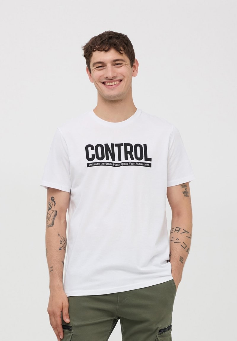 Biała bawełniana koszulka z wyraźnym czarnym nadrukiem "CONTROL", z krótkim rękawem i luźnym krojem. Noszona z oliwkowymi spodniami.