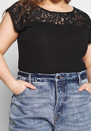 Kvinde iført sort blonder-top med kasketærmer og højtaljede acid-wash blå jeans, hænderne i lommerne, foran en ensfarvet baggrund.