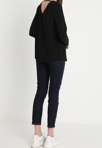 Černá blůza s dlouhým rukávem a hlubokým výstřihem na zádech, kombinovaná s tmavými džíny a bílými teniskami, která ukazuje uvolněný střih a hladkou texturu.