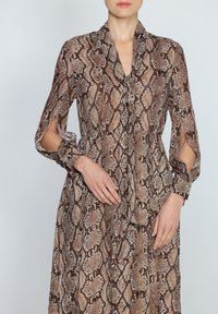 Brązowa sukienka w print węża z wiązanym kołnierzykiem, długimi rękawami z rozcięciami i dopasowanym w pasie. Wykonana z lekkiego materiału o teksturowanym wykończeniu.