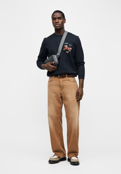 Άνδρας που φοράει μαύρο φούτερ με σχέδιο αλόγου, καφέ τζιν, ζώνη, μπεζ παπούτσια και κρατά γκρι τσάντα χιαστί, σε απλό φόντο.